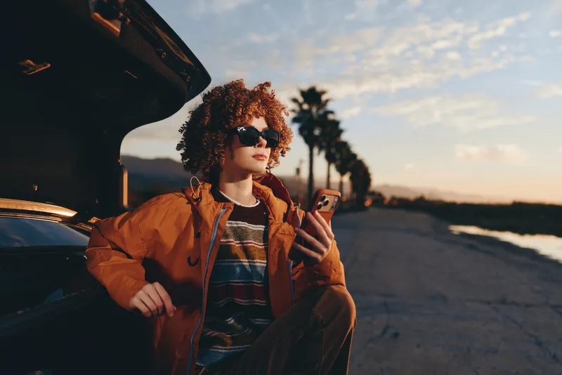 Woman lifestyle outdoors at sunset, leaning by car, wearing a jacket, curly hair, holding a phone, smiling on a road, golden light, tranquil evening bokeh