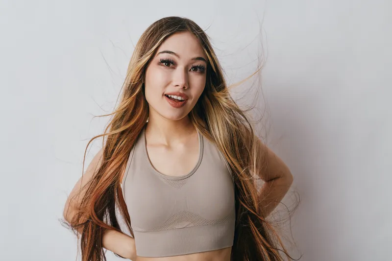 Young woman with long hair wearing sleeveless top stands smiling with hands on hips against light background. Portrait, beauty, casual and confident pose for lifestyle concept.