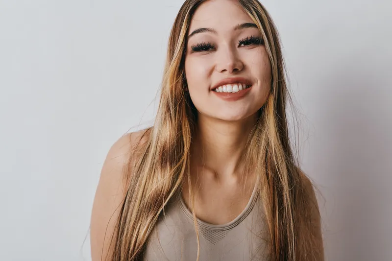 Smiling young woman with long hair wearing casual top posing against plain wall, portrait with natural light and friendly expression for lifestyle and beauty concept.