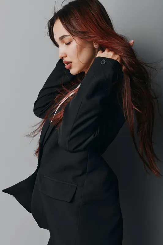 Woman with long hair wearing black blazer posing sideways with eyes closed in studio. Fashion model with natural makeup and stylish outfit on gray background for beauty and style.