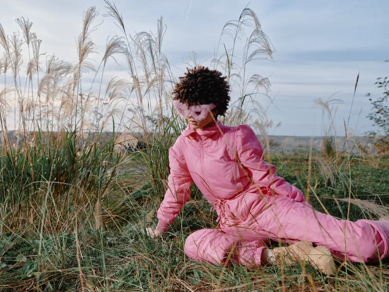 Outdoor person in a pink jumpsuit performs a wide stretch amid tall grasses on an open field, blue sky above, capturing movement, balance and natural calm.