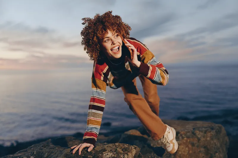 Woman lifestyle at seaside ocean, phone call by ear, curly hair, smiling outdoors, standing on rocky coastline, casual jacket and scarf, windy atmosphere, dramatic clouds