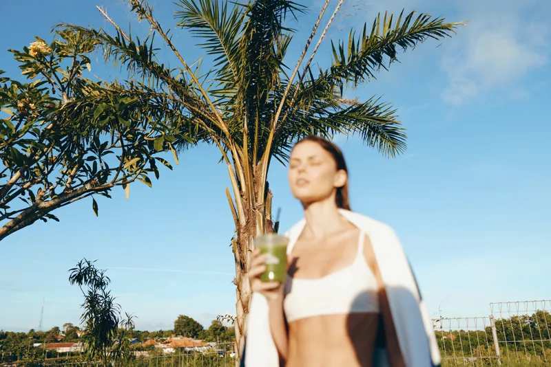 Young woman in white sportswear holding green smoothie outdoors near palm tree, sunny day, healthy lifestyle, relaxed mood, natural environment, summer scene