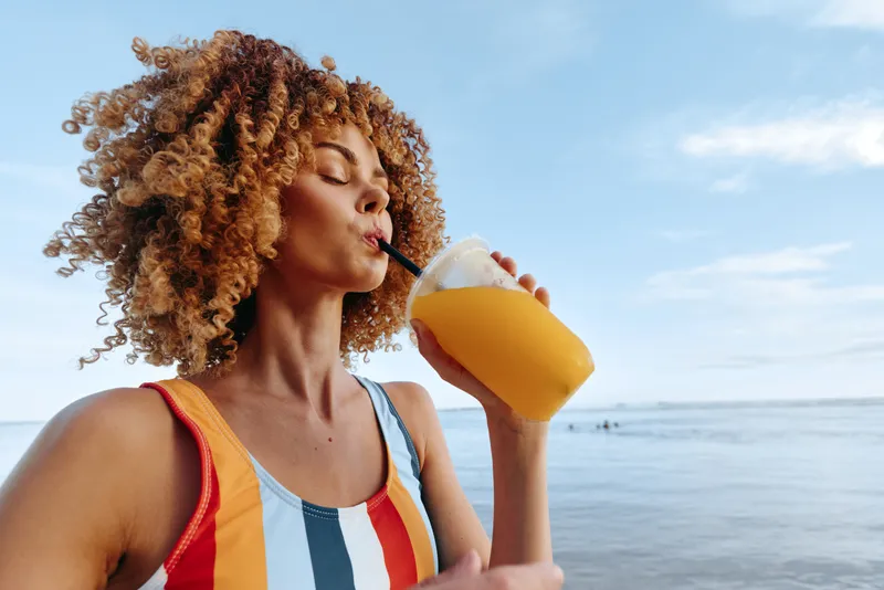 Woman with curly hair drinking juice on a beach, summer hydration concept, outdoor relaxation in bright daylight, healthy lifestyle, refreshing drink, side view against blue sky
