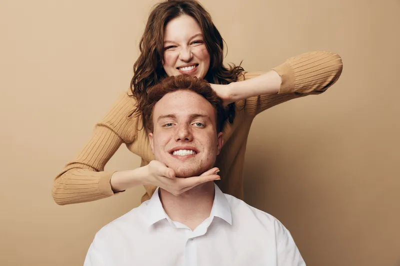 Smiling young woman playfully holds a young man’s head with her hands. Both have happy expressions, casual clothing, and stand against a plain beige background. Fun friendship concept.