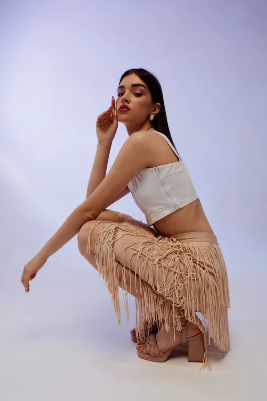 Young woman in stylish beige fringed pants and white crop top poses in studio. Concept for woman beauty fashion with modern outfit and confident attitude.