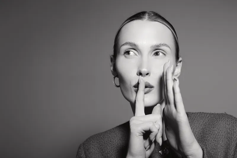 A young white woman with light skin and blonde hair gestures silence with finger on lips. Black and white portrait with serious expression on grey background.