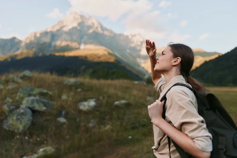 Cheerful woman hiking outdoors, backpacked traveler, sunlit mountain landscape, relaxed posture, fresh breeze, natural colors, expansive scenery, sense of adventure, open sky