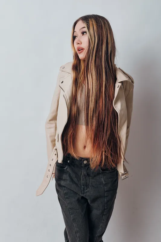 Young woman with long straight hair wearing light jacket and black jeans posing against plain white background for fashion and style portrait.