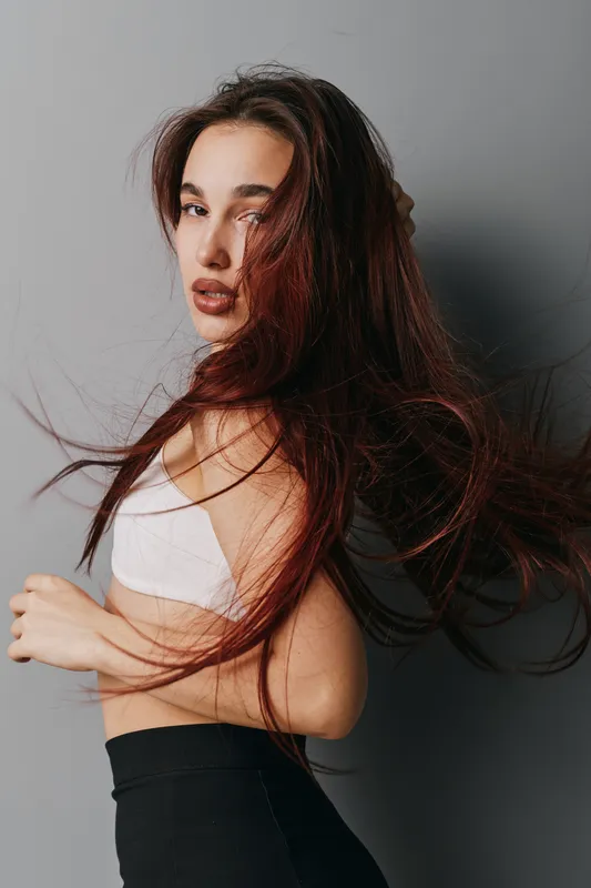 Woman with long flowing hair posing in white top and black pants against gray background. Portrait of confident female with natural makeup and dynamic hairstyle in studio setting.