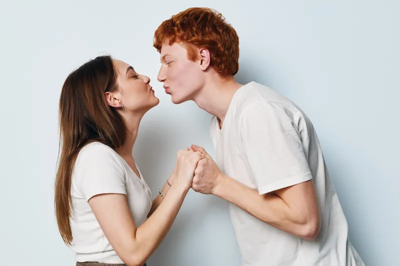 Young couple holding hands and leaning forward for a kiss in casual white t shirts with closed eyes in studio on light background. Love, romance and connection between man and woman.
