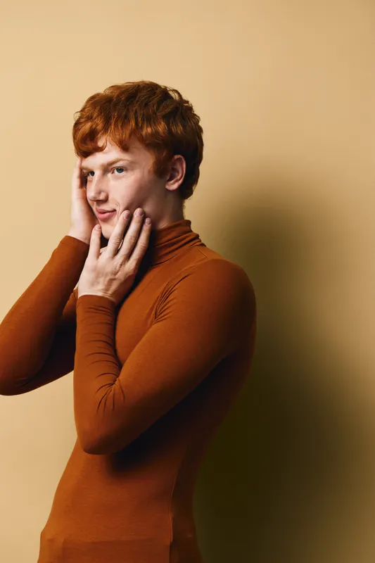 Portrait of a young redhead man with curly hair wearing a brown turtleneck posing with hands on face against beige background. Expression, profile and fashion style in studio lighting.