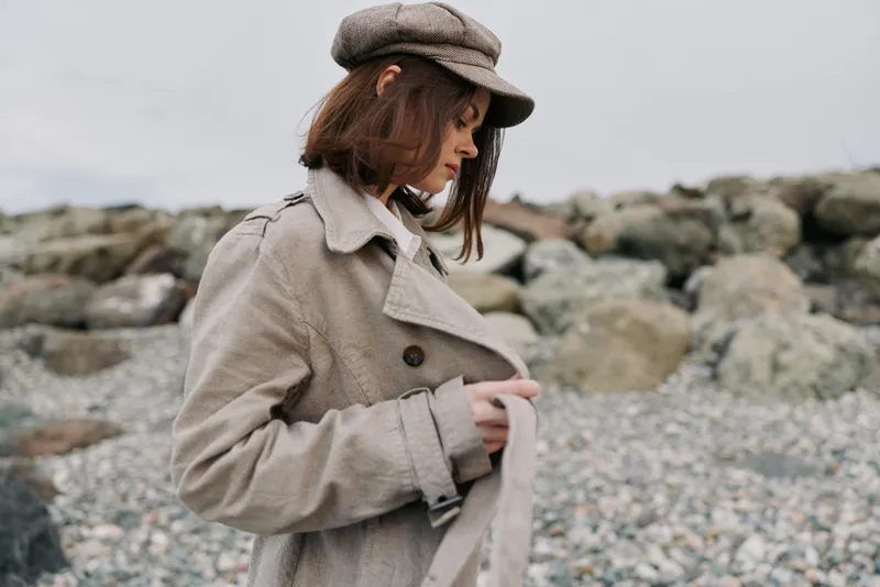 Young woman in beige trench coat and hat stands on rocky beach looking down, thoughtful mood. Overcast sky and natural stones create calm, introspective atmosphere outdoors.