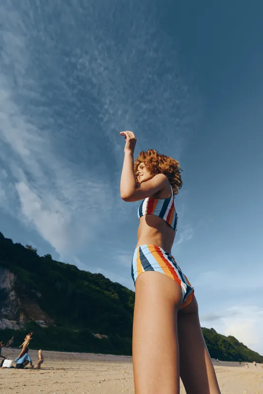 Young woman in colorful striped swimsuit standing on sandy beach, looking up with hand raised, clear blue sky, green hills background, summer outdoor scene