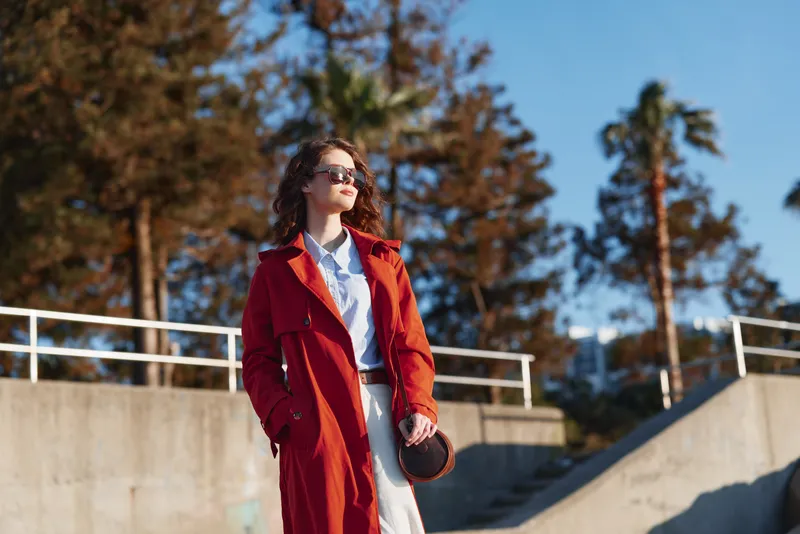 Young woman in red trench coat and sunglasses walks confidently outdoors on a sunny autumn day with trees and concrete stairs in the background.