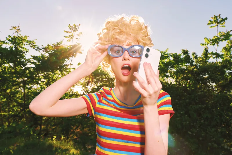 A young woman in a bright striped shirt poses outdoors, wearing retro sunglasses and filming a selfie with a phone. Retro film color captures a carefree summer moment.
