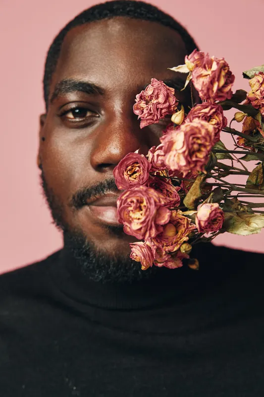 African man with dark skin and beard holding dried pink flowers near face in creative conceptual visual style on pink background. Portrait with artistic expression and floral elements.
