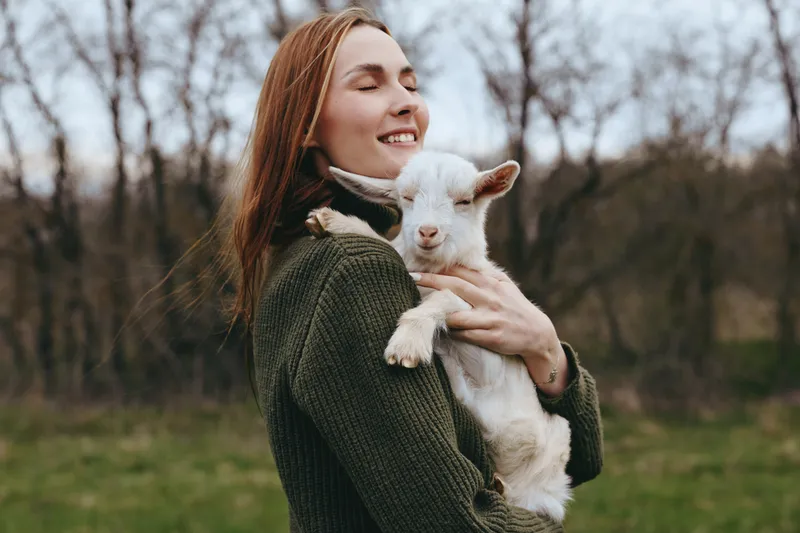 Joyful woman outdoors hugs a white goat kid, expressing warm affection in a rustic setting, showcasing countryside lifestyle and companionship with animals, natural light, candid moment, peaceful