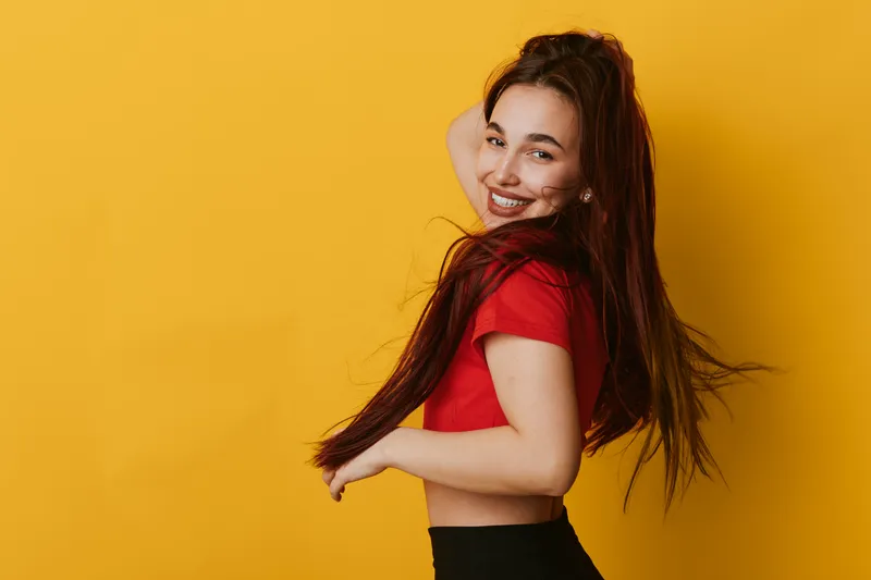 Young woman with long hair wearing red shirt smiling and posing on yellow background. Female model with natural makeup in casual outfit showing joyful expression and confidence.