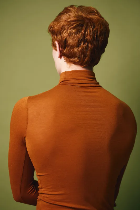 Redheaded man with short curly hair wearing tight orange turtleneck sweater standing with back to camera against green background indoors.