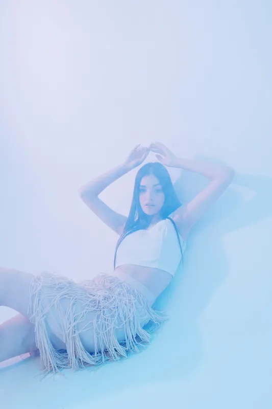 Young woman in a white crop top and fringed skirt poses against a light blue background. The image focuses on beauty, fashion, and modern style concepts.