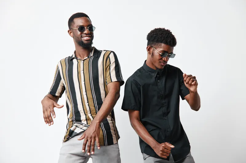 Two young black men with dark skin dancing and smiling in casual summer clothes and sunglasses on white background. Friends enjoying music and having fun together indoors.