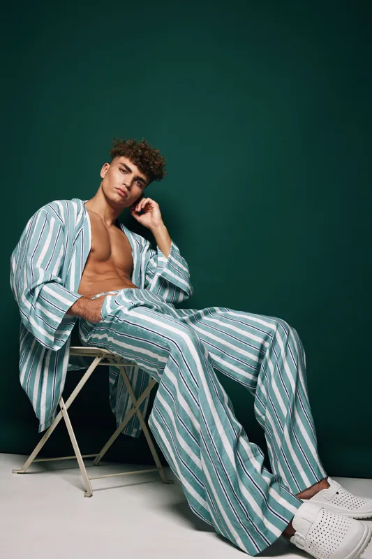 Young man wearing loose striped shirt and pants posing on chair against green background. Fashion model with bare chest and curly hair sitting barefoot in casual clothing style.