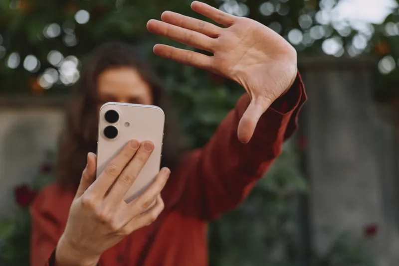 Lifestyle portrait of a woman in a red top taking a selfie with a smartphone, hand reaching toward the lens, outdoors with soft blurred background and natural light