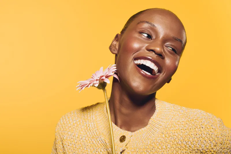 Happy African woman with dark skin laughing while holding a pink flower near her face on yellow background. Concept of joy, beauty, and natural skincare for black female model.