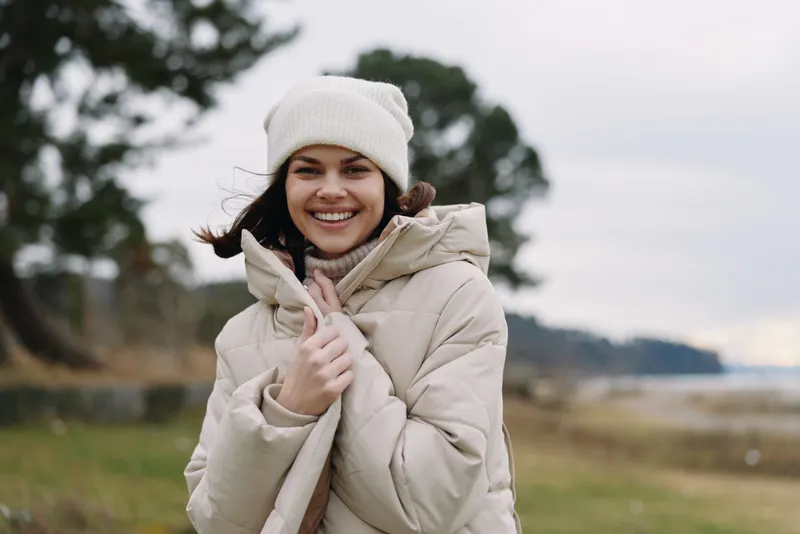 Woman in winter clothing and knit hat smiles outdoors, thumbs up, standing in a chilly landscape with water and trees, conveying warmth, wellness and outdoor joy.