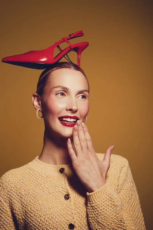 Smiling woman with red high heel shoe on her head wears yellow knitted sweater, posing on brown background. Concept of fashion, creativity, and playful style in studio lighting.