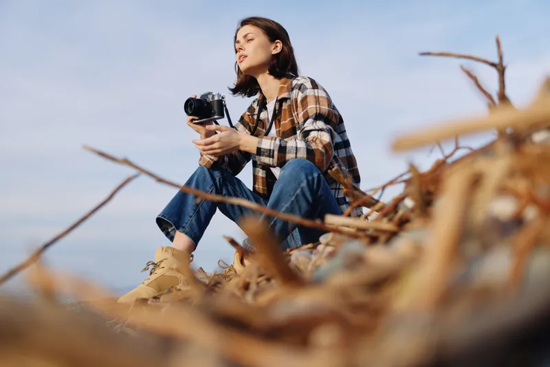 Photographer with camera in hand in outdoors scene, a woman wearing plaid shirt among driftwood, capturing nature and landscape with focused, artistic composition.