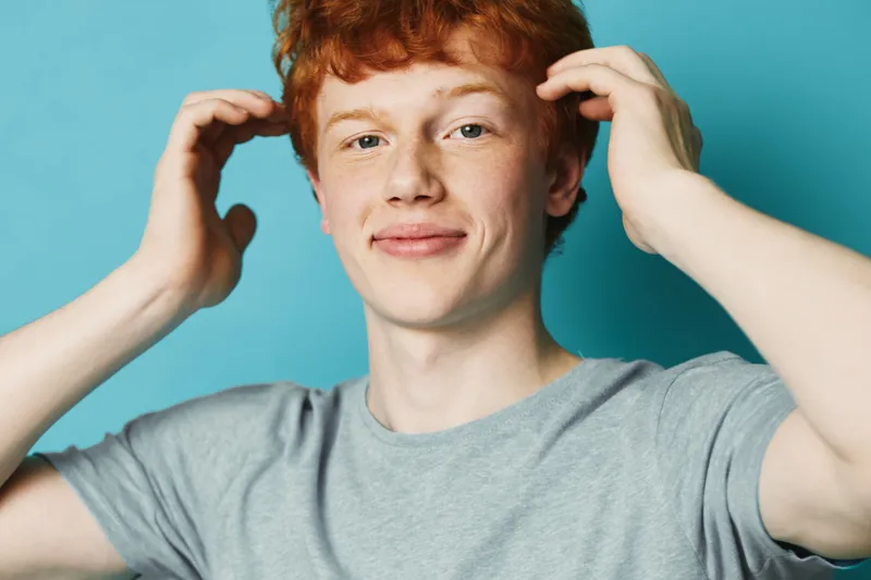 Young man with red hair and freckles wearing gray t shirt posing and smiling on blue background, casual style and natural look with playful gesture by hands near head.