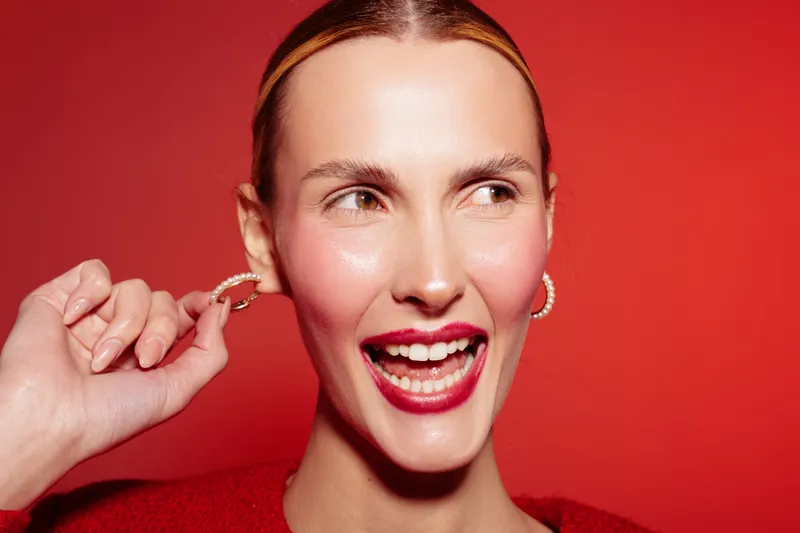 Close-up of smiling woman with pearl earrings and red lipstick on red background. Concept of beauty, fashion, makeup, and joyful expression in studio lighting.