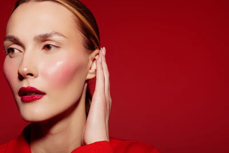Close-up of a woman with red lipstick and glowing skin posing against a red background. Concept of beauty, makeup, skincare, and fashion with a confident expression.