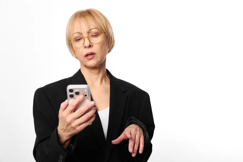 Woman businesswoman holding smartphone, phone in hand, focused expression, office blazer on white background, communication gesture, portrait view, natural light, minimal setting