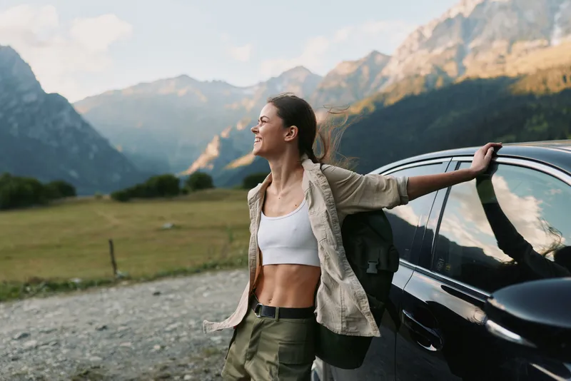 A woman stands beside a car in a wide meadow, arms outstretched, savoring freedom and nature as mountains rise behind, capturing a carefree, outdoors lifestyle moment