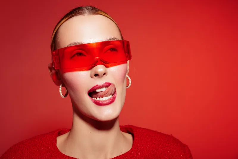Young woman wearing red futuristic glasses and red sweater poses with playful expression on red background. Concept of fashion, style, modern accessories, and vibrant portrait.