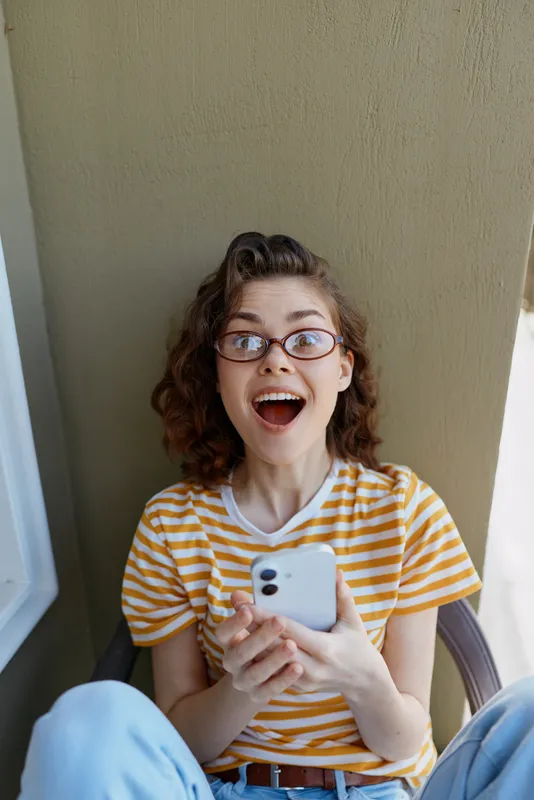Young woman with curly hair and glasses enjoys a candid moment on a balcony, wearing a yellow striped t shirt, holding a smartphone and reacting with joyful surprise