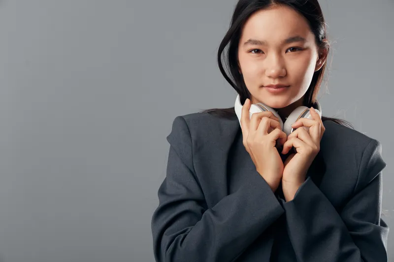 Young woman with headphones holding them close to face against gray background. Portrait of confident female in dark jacket looking at camera, modern style with natural expression and minimal makeup.