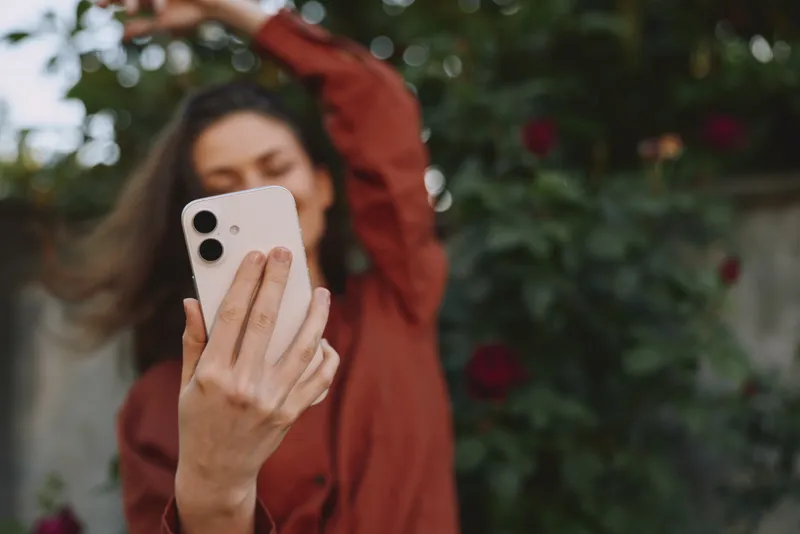 Young woman takes a selfie in a garden with roses, wearing a maroon top, smiling softly at the phone, capturing a casual outdoor moment