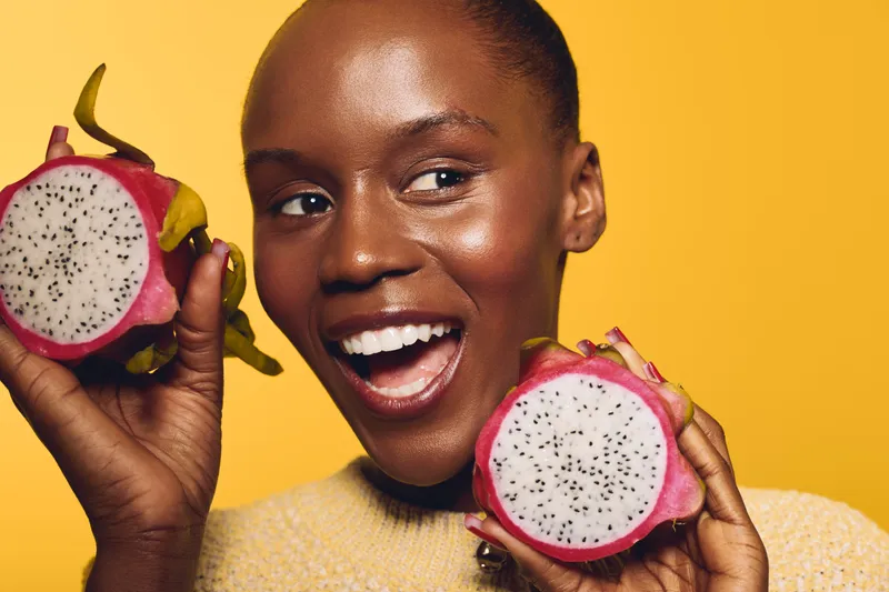 Happy African woman with dark skin holds two halves of dragon fruit close to her face, smiling with open mouth on yellow background. Concept of healthy eating, exotic fruit, and joy.