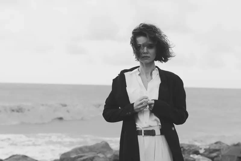 Woman in a tailored coat and white shirt stands by the sea in a windy, gray shoreline scene. Monochrome photography emphasizes fashion, movement, and a moody, contemplative atmosphere