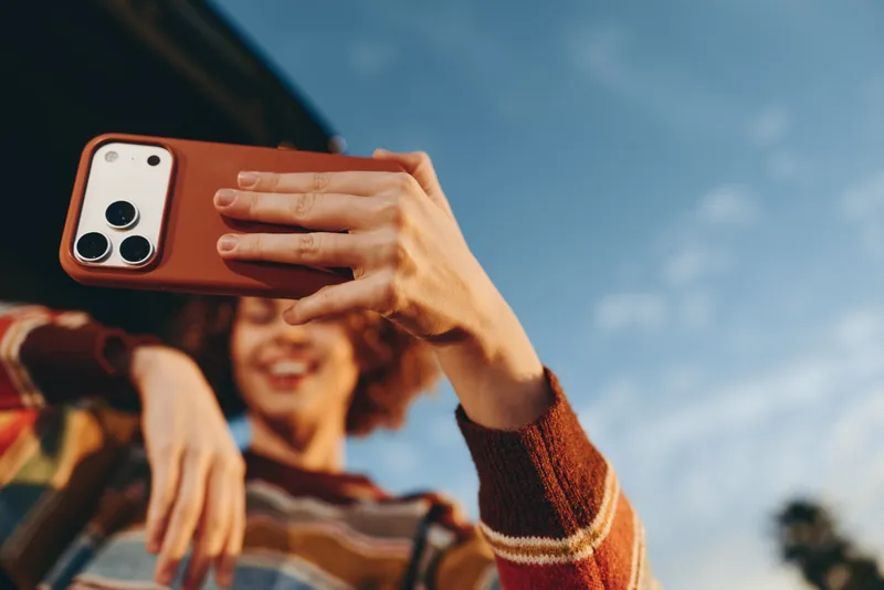 Woman outdoors taking a selfie with smartphone, smiling and laughing in sunlit moment, casual travel lifestyle, relaxed hand gesture, clear sky, blurred background, bright colors, warm smile