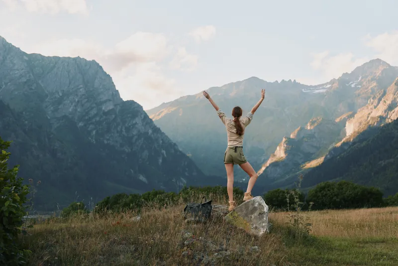 A woman stands on a rock with arms raised in a bright mountain landscape, embodying achievement, freedom, and joy in outdoor adventure, with distant peaks and sunlit skies.