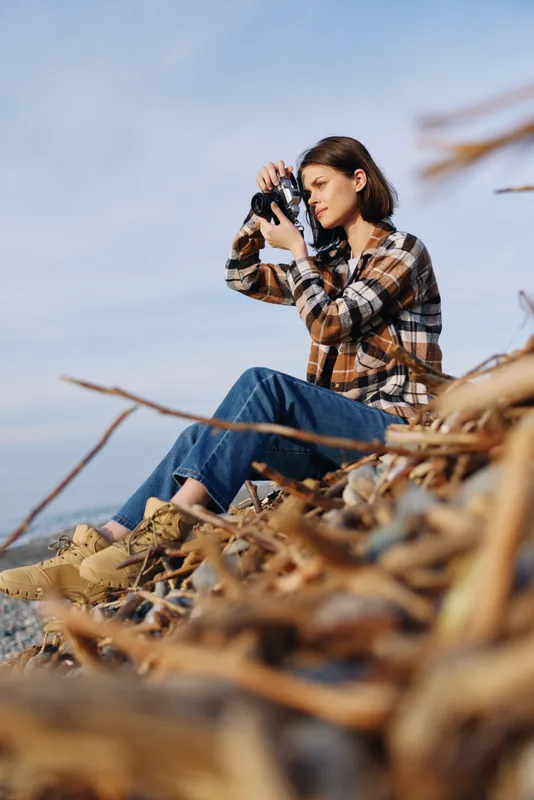 Photographer outdoors with camera on a rocky shore, focused on distant landscape and autumn scenery, capturing nature's calm with warm light and breeze
