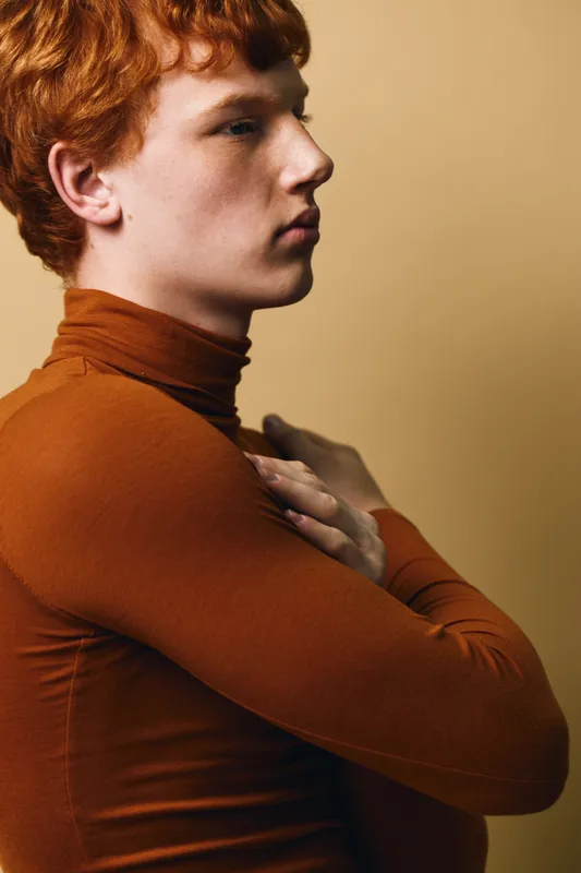 Young man with red hair and freckles in brown turtleneck sweater posing in profile on neutral background. Thoughtful expression with hand on chest and soft lighting.