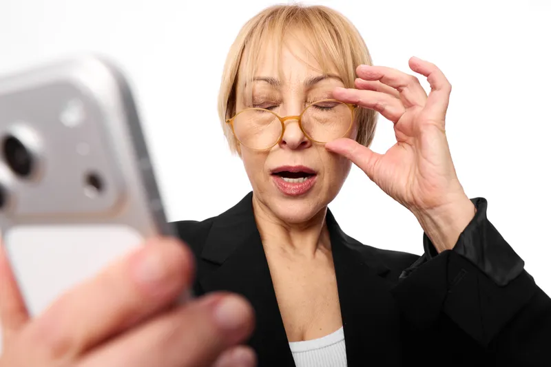 Elderly woman, business attire, holding glasses and touching face, eyes closed, eyesight strained, tired stress, smartphone in foreground, white background, expression