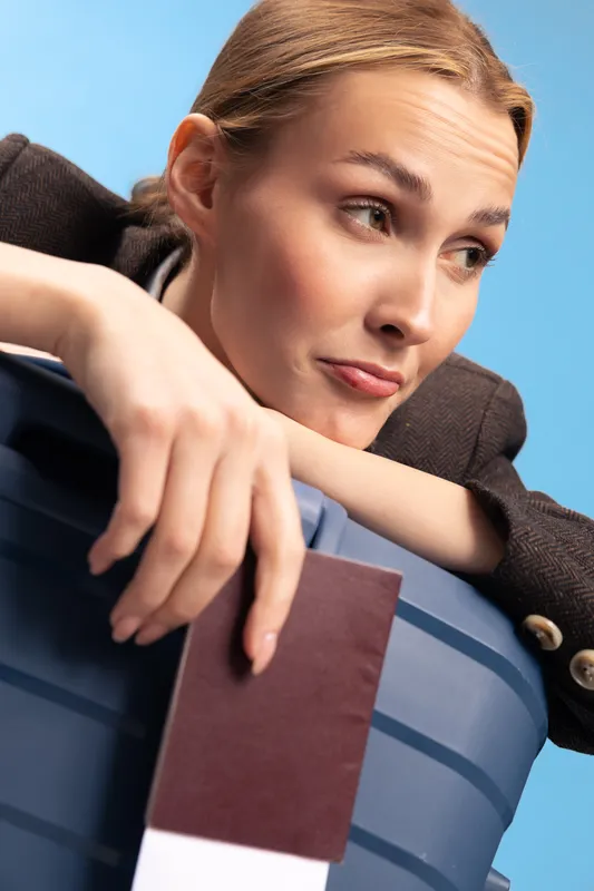 Young woman leans on suitcase holding a passport and ticket with thoughtful expression. Travel concept, preparation, and planning for trip or vacation on blue background.