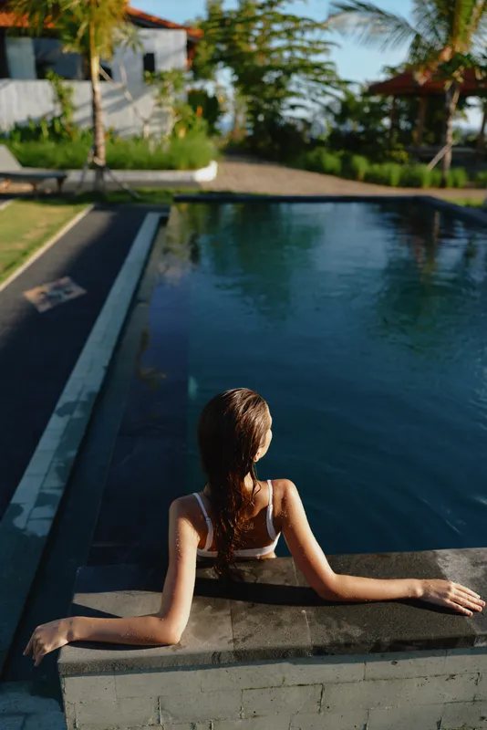A woman relaxes by a sunny pool, leaning on the edge with her back to the camera. A candid lifestyle moment captures warm light, shimmering water, and a peaceful outdoor retreat.